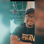 A man wearing glasses, a black cap, and a green Vans t-shirt smiles while sitting at an outdoor covered patio, enjoying the day after visiting top pet groomers in NYC and Queens.