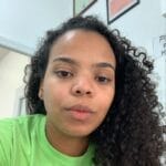 A woman with curly hair wearing a light green shirt looks at the camera in an indoor setting, with framed certificates—similar to those found in top pet groomers NYC Queens NJ—on the wall behind her.