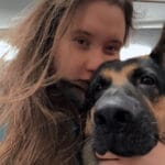 A woman with long brown hair holds her German Shepherd close for a selfie indoors under fluorescent lighting, showing the special bond found at trusted pet groomers in NYC, Queens, or NJ.